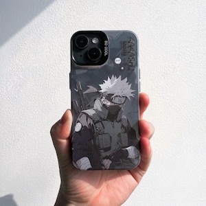 May include: A phone case featuring a detailed anime illustration of a character in shades of gray and black. The case has the text "KAKASHI NARUTO" and "SO COOL" printed on it. The case is held in a hand.
