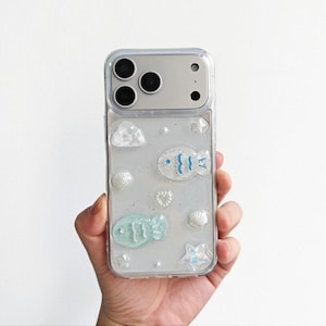 Könnte beinhalten: Transparente Handyhülle mit maritimem Thema. Sie zeigt blaue und weiße Fische, Muscheln, Wolken und sternförmige Verzierungen. Die Hülle ist für ein Smartphone mit mehreren Kameralinsen konzipiert.