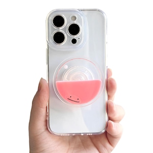 May include: A clear smartphone case featuring a circular, collapsible phone grip attached to the back. The grip has a semi-circular pink silicone top with a simple black line representing a mouth, resembling a stylized character face.