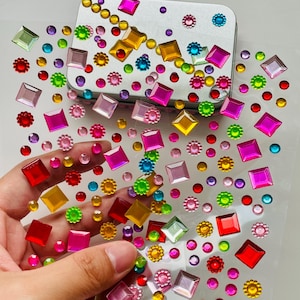 May include: A clear adhesive sheet covered in a variety of colorful plastic rhinestones in shapes including squares, circles, and flowers. The gems feature a vibrant palette of red, pink, yellow, green, blue, and purple, arranged in a scattered pattern.