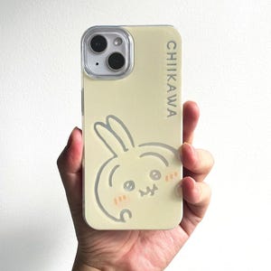以下が含まれることがあります： 大きな目と小さなピンクの頬をしたウサギのキャラクターのグラフィックが型押しされた、淡い黄色のスマートフォンケース。ケースの右側には、CHIIKAWAという文字がダークグレーのサンセリフ体で縦書きにプリントされています。