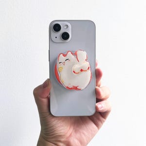 Könnte beinhalten: Ein weiß-oranger Katzen-Handyhalter ist an einem silbernen Smartphone befestigt. Die Katze hat ein lächelndes Gesicht und ist von einem roten Rand umgeben. Das Telefon wird in einer Hand vor weißem Hintergrund gehalten.