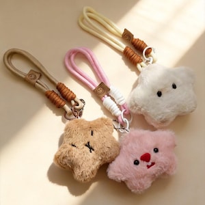 May include: Three plush star-shaped keychains with soft faux fur. One is brown with stitched eyes and mouth, one is pink with embroidered facial features, and one is white with black eyes. Each has a looped wrist strap with a small smiley face tag.