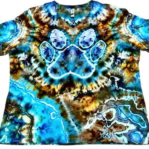 May include: A short-sleeve V-neck t-shirt featuring a tie-dye pattern in shades of bright blue, dark brown, and white. The center of the shirt displays a prominent paw print graphic outlined in white, surrounded by swirling, marbled color effects.
