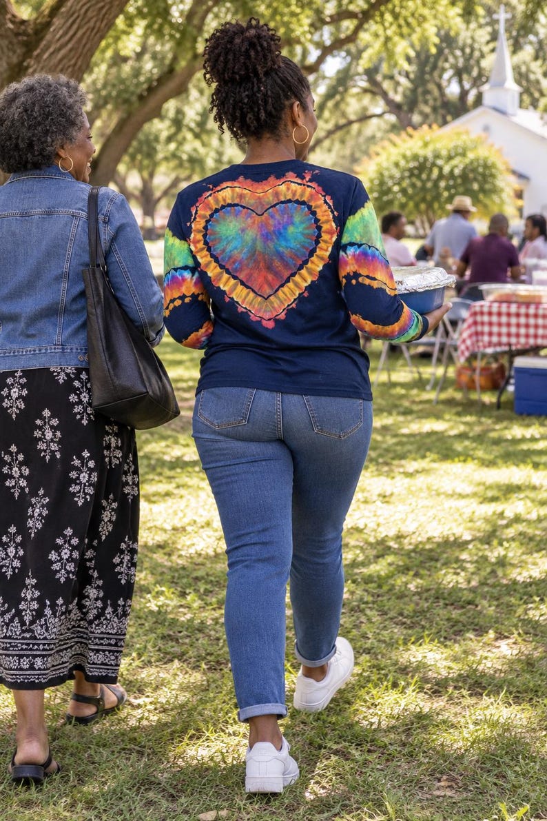 May include: A long-sleeved navy blue shirt featuring a large, vibrant tie-dye heart graphic on the back, paired with blue denim jeans and white trainers. The shirt sleeves also display matching tie-dye patterns in bright rainbow colours.