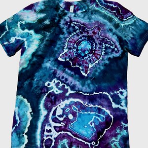 May include: A short-sleeve crew neck t-shirt featuring a tie-dye pattern in shades of deep purple, navy blue, and bright turquoise. The design includes a prominent central graphic of a sea turtle surrounded by abstract, swirling geode-like shapes.