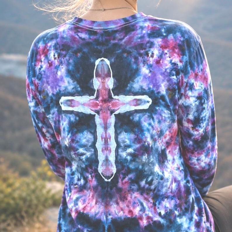 May include: A long-sleeve shirt featuring a tie-dye pattern in shades of deep blue, purple, and magenta. The center of the back displays a prominent, white, cross-shaped design created through the tie-dye process, contrasting with the dark colors.
