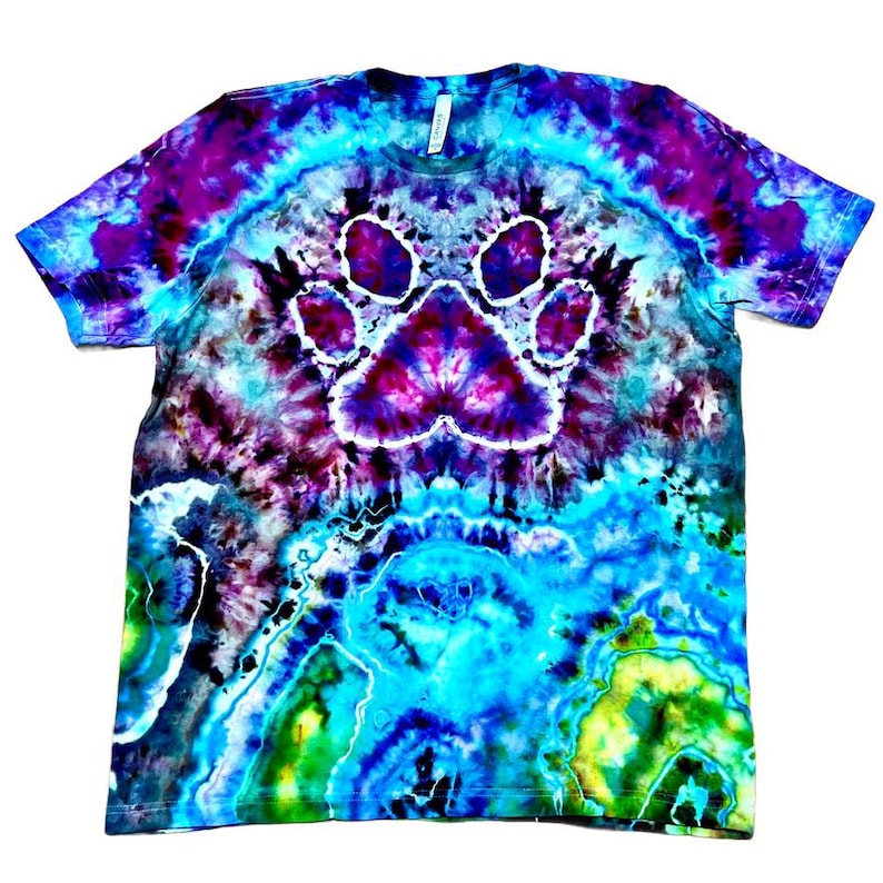 Op de afbeelding: Een tie-dye t-shirt met een groot pootafdrukontwerp in het midden. Het shirt heeft een levendige mix van kleuren, waaronder blauw, paars en groen. De pootafdruk is wit en het shirt heeft een ronde hals.