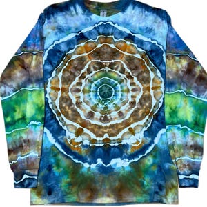 May include: A long-sleeve crew neck shirt featuring a tie-dye pattern with a central circular mandala design. The colour palette includes shades of deep blue, earthy brown, and vibrant green, separated by thin, irregular white lines throughout.