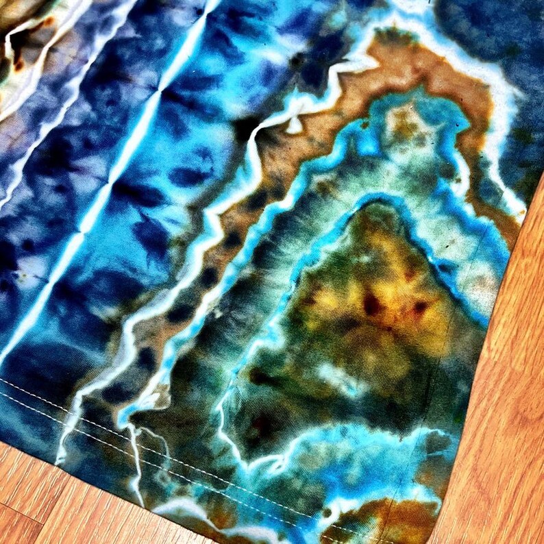 May include: Close-up of a tie-dye fabric with a vibrant mix of blues, browns, and yellows. The design features diagonal stripes and a textured appearance, with white stitching along the edge. The fabric has a unique, artistic look.