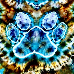 May include: A tie-dye t-shirt featuring a prominent paw print graphic centered on the fabric. The design showcases vibrant shades of blue, turquoise, and deep brown, with white outlines defining the paw shape against the swirling tie-dye pattern.