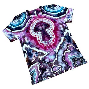 Puede incluir: Una camiseta de manga corta con un patr&oacute;n tie-dye inspirado en geodas en tonos magenta intenso, morado oscuro, negro y turquesa brillante. El dise&ntilde;o incluye formas circulares y org&aacute;nicas conc&eacute;ntricas con contornos blancos en la tela.