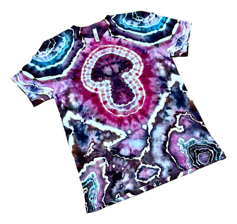 Puede incluir: Una camiseta de manga corta con un patr&oacute;n tie-dye inspirado en geodas en tonos magenta intenso, morado oscuro, negro y turquesa brillante. El dise&ntilde;o incluye formas circulares y org&aacute;nicas conc&eacute;ntricas con contornos blancos en la tela.