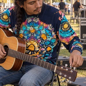 May include: A long-sleeve shirt featuring a dark navy base with a vibrant tie-dye pattern in shades of yellow, orange, pink, and turquoise. The design includes circular motifs across the chest and sleeves, paired with blue denim jeans.