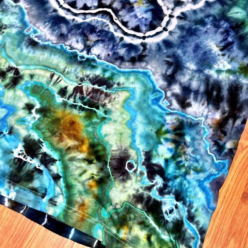 May include: Close-up of a tie-dye fabric with a blend of blues, greens, and yellows. The design features organic, flowing patterns with white accents, creating a marbled effect. The fabric's edge is visible, showing a hem.