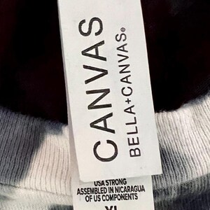 May include: Close-up of a white tag attached to a gray and white tie-dye t-shirt. The tag reads "CANVAS" and "BELLA + CANVAS." Below the brand names, the tag states "USA STRONG, ASSEMBLED IN NICARAGUA OF US COMPONENTS" and the size "XL."