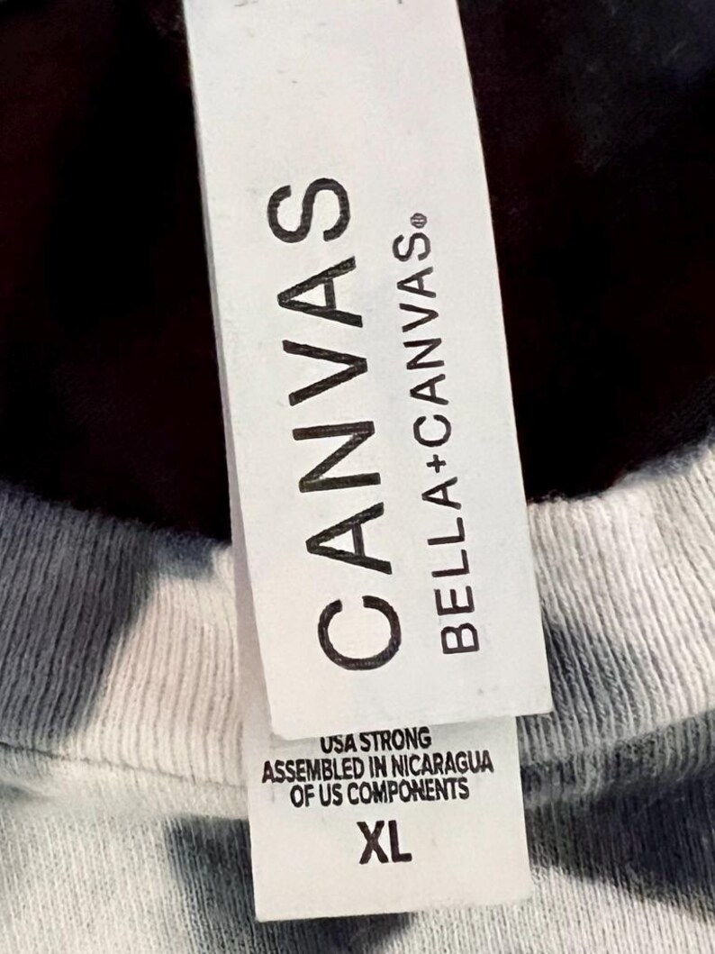 May include: Close-up of a white tag attached to a gray and white tie-dye t-shirt. The tag reads "CANVAS" and "BELLA + CANVAS." Below the brand names, the tag states "USA STRONG, ASSEMBLED IN NICARAGUA OF US COMPONENTS" and the size "XL."