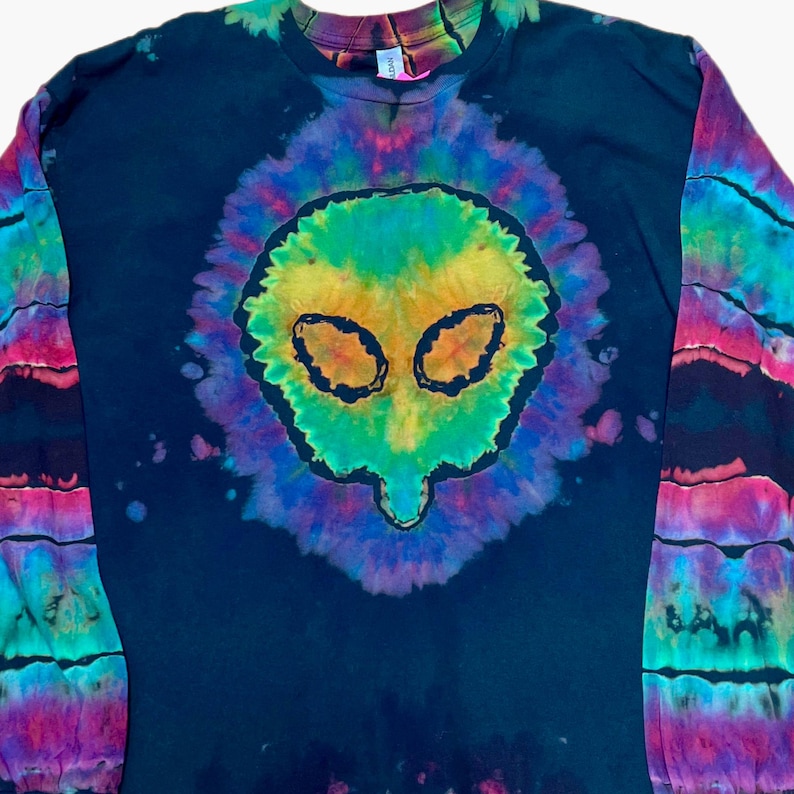 May include: A long-sleeve black t-shirt featuring a central tie-dye alien head graphic in vibrant shades of green, yellow, and purple. The sleeves display horizontal bands of tie-dye patterns in pink, blue, and green, creating a bold contrast.