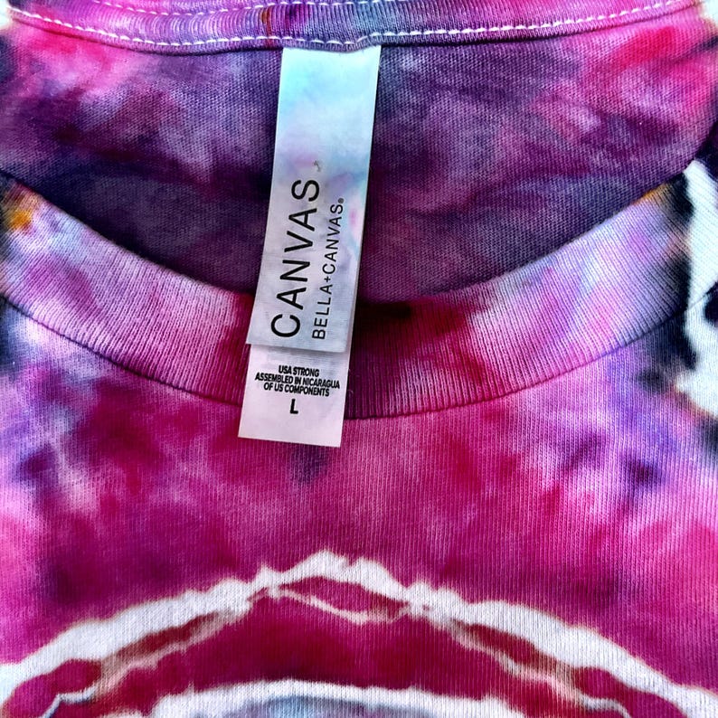 Puede incluir: Una camiseta tie-dye con una mezcla vibrante de colores rosa, morado y negro. La prenda incluye una etiqueta blanca en el cuello con el texto CANVAS BELLA+CANVAS, USA STRONG, ASSEMBLED IN NICARAGUA OF US COMPONENTS y la letra de talla L.