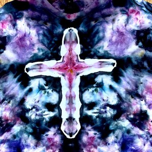 May include: A short-sleeve t-shirt featuring a tie-dye pattern in shades of black, deep purple, and blue. The center of the garment displays a prominent white cross shape with pink and purple accents, created through a tie-dye technique.