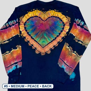 May include: A long-sleeve black shirt featuring a large, central tie-dye heart pattern in shades of yellow, orange, pink, purple, and blue. The sleeves have matching tie-dye stripes. A white label at the bottom reads #5 &bull; MEDIUM &bull; PEACE &bull; BACK.