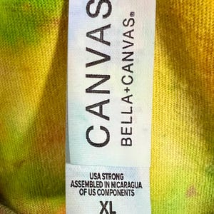 Może przedstawiać: Zbliżenie na metkę koszulki tie-dye. Na metce widnieje napis "CANVAS BELLA+CANVAS". Poniżej nazwy marki widnieje napis "USA STRONG ASSEMBLED IN NICARAGUA OF US COMPONENTS". Rozmiar to XL. Koszulka ma kolorowy, mieszany wz&oacute;r.