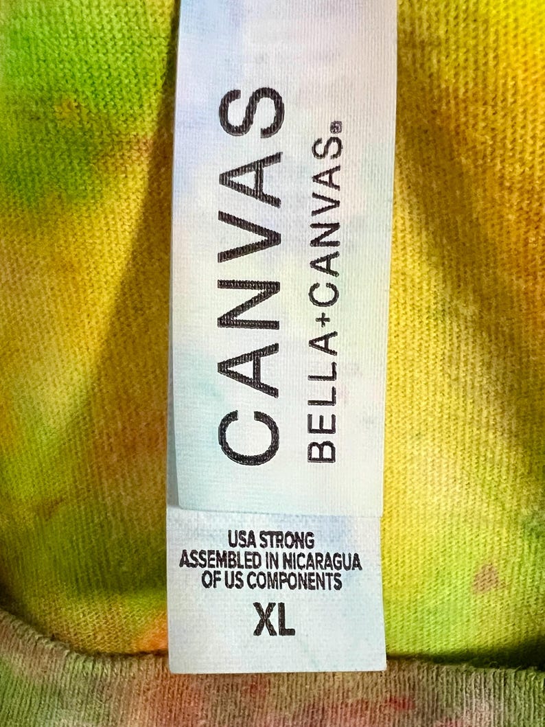 Może przedstawiać: Zbliżenie na metkę koszulki tie-dye. Na metce widnieje napis "CANVAS BELLA+CANVAS". Poniżej nazwy marki widnieje napis "USA STRONG ASSEMBLED IN NICARAGUA OF US COMPONENTS". Rozmiar to XL. Koszulka ma kolorowy, mieszany wz&oacute;r.