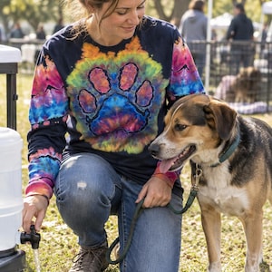 May include: A long-sleeve shirt featuring a dark navy base with a vibrant, multi-colored tie-dye pattern. The center of the shirt displays a large, bold paw print graphic in shades of yellow, orange, pink, and blue against the dark fabric.