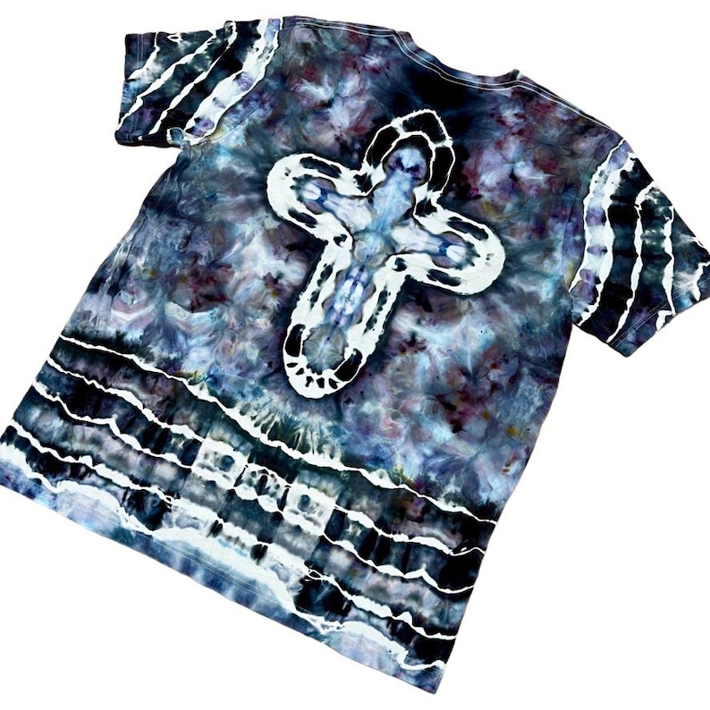 May include: Tie-dye t-shirt with a large white cross design on the back. The shirt features a blend of dark blue, purple, and black colors with white accents. The sleeves also have white stripes.