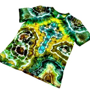 Puede incluir: Una camiseta te&ntilde;ida con tinte en tonos verdes, amarillos, marrones y azules. La camiseta presenta un dise&ntilde;o de cruz en el centro y un patr&oacute;n &uacute;nico y arremolinado. La camiseta de manga corta est&aacute; hecha de tela suave.
