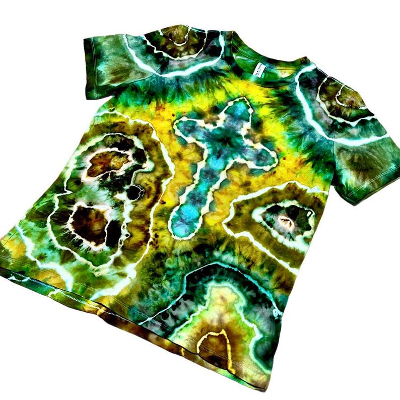 Puede incluir: Una camiseta te&ntilde;ida con tinte en tonos verdes, amarillos, marrones y azules. La camiseta presenta un dise&ntilde;o de cruz en el centro y un patr&oacute;n &uacute;nico y arremolinado. La camiseta de manga corta est&aacute; hecha de tela suave.