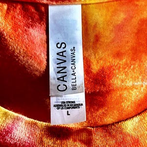 May include: Close-up of a tie-dye t-shirt with a tag that reads "CANVAS BELLA+CANVAS." The tag also states "USA STRONG ASSEMBLED IN NICARAGUA OF US COMPONENTS" and has a size "L". The shirt has a blend of orange, yellow, and red colours.
