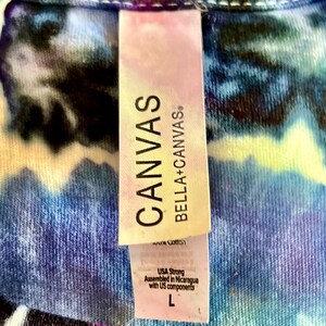 以下が含まれることがあります： タイダイ柄のシャツのクローズアップ。タグには「CANVAS BELLA+CANVAS」と書かれています。タグには、このシャツはUSA Strongで、ニカラグアで米国のコンポーネントを使用して組み立てられ、サイズLであることも記載されています。