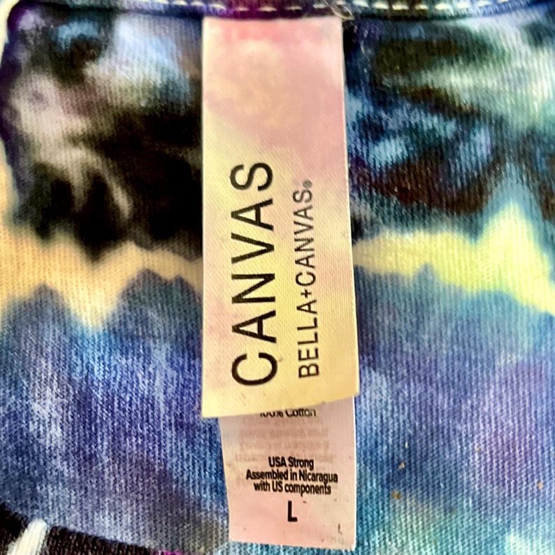 以下が含まれることがあります： タイダイ柄のシャツのクローズアップ。タグには「CANVAS BELLA+CANVAS」と書かれています。タグには、このシャツはUSA Strongで、ニカラグアで米国のコンポーネントを使用して組み立てられ、サイズLであることも記載されています。