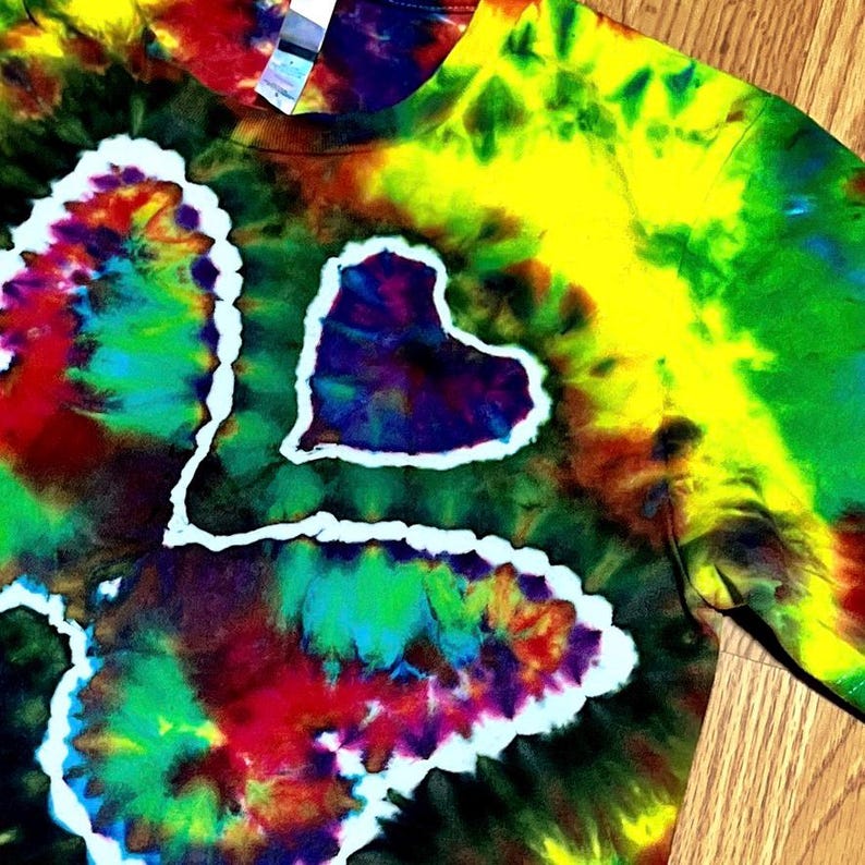 May include: A tie-dye t-shirt with a heart design. The shirt features a vibrant mix of colors, including yellow, green, red, and blue, creating a psychedelic effect. The heart shape is outlined in white, adding contrast to the colorful background.