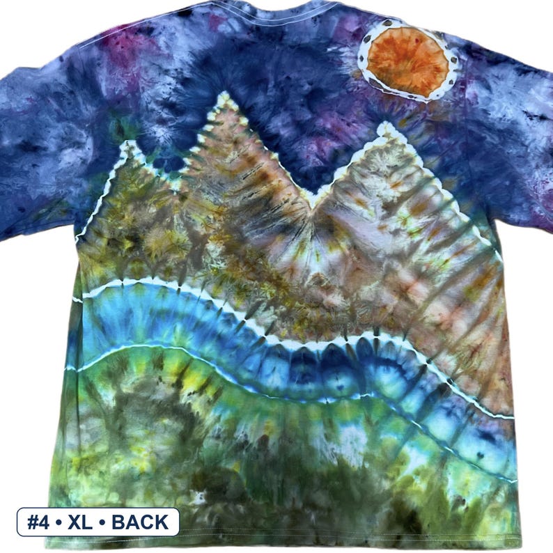 May include: A short-sleeve t-shirt featuring a tie-dye mountain landscape design. The scene includes a dark blue sky with an orange sun, brown mountain peaks, and a blue and green river flowing through the foreground. Text reads #4 &bull; XL &bull; BACK.