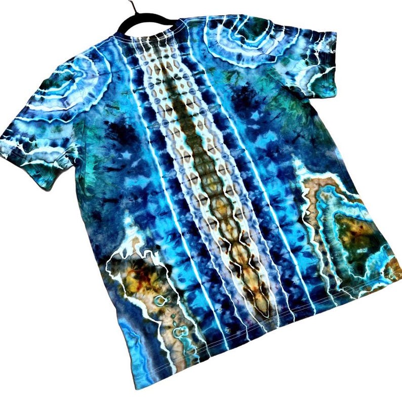 May include: A short-sleeved tie-dye t-shirt in shades of blue, teal, and brown. The shirt features a central vertical pattern with white and brown accents, and a swirling design on the sleeves.