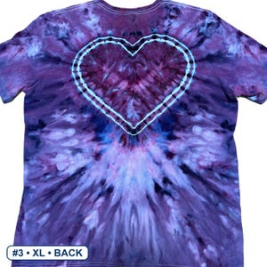 Może przedstawiać: Koszulka z kr&oacute;tkim rękawem z wzorem tie-dye w odcieniach fioletu, błękitu i bieli. Wz&oacute;r zawiera wyraźny kształt serca umieszczony na środku plec&oacute;w, obrysowany białą linią. Etykieta w lewym dolnym rogu zawiera napis #3 &bull; XL &bull; BACK.