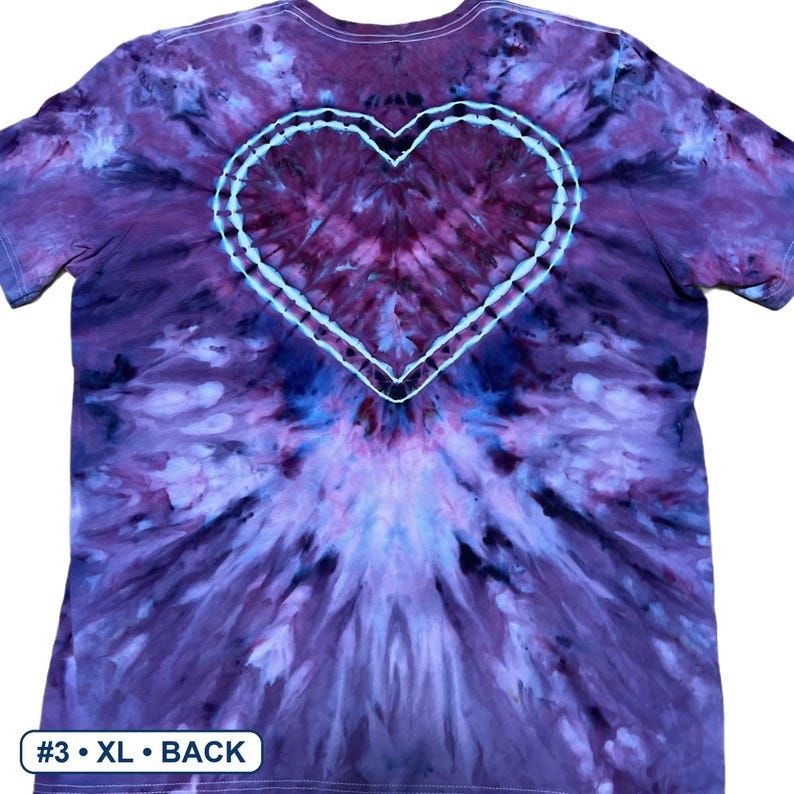Może przedstawiać: Koszulka z kr&oacute;tkim rękawem z wzorem tie-dye w odcieniach fioletu, błękitu i bieli. Wz&oacute;r zawiera wyraźny kształt serca umieszczony na środku plec&oacute;w, obrysowany białą linią. Etykieta w lewym dolnym rogu zawiera napis #3 &bull; XL &bull; BACK.