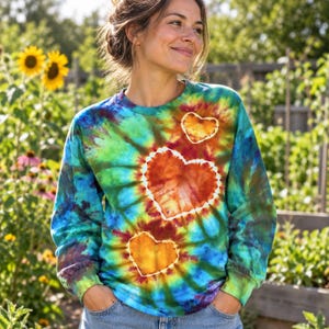 Może przedstawiać: Bluza z długim rękawem i okrągłym dekoltem, ozdobiona żywym wzorem tie-dye w odcieniach niebieskiego, zielonego i ż&oacute;łtego. Z przodu znajdują się trzy wyraźne kształty serc w kolorze pomarańczowym i czerwonym, każde z białą obw&oacute;dką na tle tie-dye.