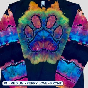 May include: A black long-sleeve shirt featuring a central tie-dye paw print design in vibrant shades of yellow, orange, pink, purple, and blue. The sleeves also display horizontal tie-dye bands. Text at the bottom reads #1 &bull; MEDIUM &bull; PUPPY LOVE &bull; FRONT.
