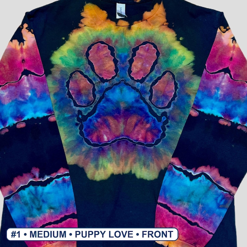 May include: A black long-sleeve shirt featuring a central tie-dye paw print design in vibrant shades of yellow, orange, pink, purple, and blue. The sleeves also display horizontal tie-dye bands. Text at the bottom reads #1 &bull; MEDIUM &bull; PUPPY LOVE &bull; FRONT.