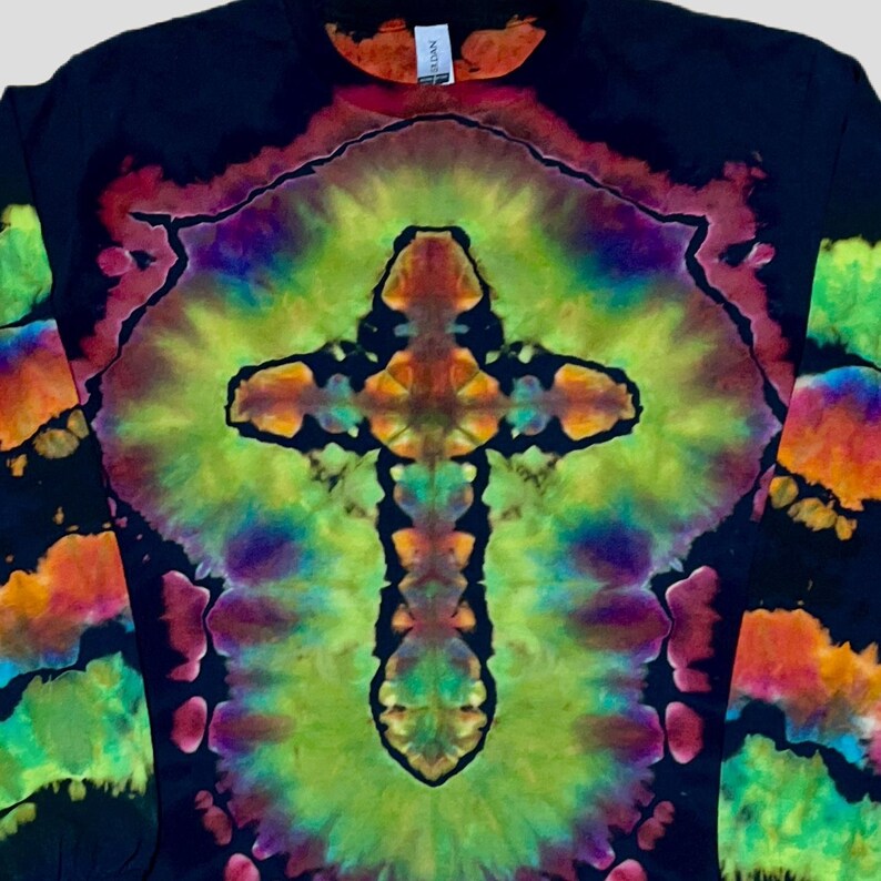 May include: A long-sleeve shirt featuring a black base with a vibrant tie-dye pattern. The centre displays a prominent cross shape in shades of lime green, orange, and purple, surrounded by a radiating halo effect. The sleeves also feature tie-dye.