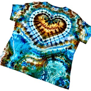 May include: A short-sleeve t-shirt featuring a tie-dye pattern in shades of turquoise, deep blue, brown, and black. The design showcases a prominent central heart shape outlined in white, surrounded by intricate, swirling geode-like patterns.