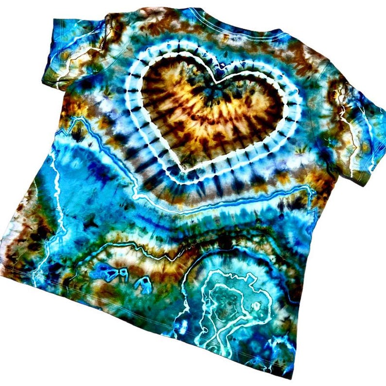 May include: A short-sleeve t-shirt featuring a tie-dye pattern in shades of turquoise, deep blue, brown, and black. The design showcases a prominent central heart shape outlined in white, surrounded by intricate, swirling geode-like patterns.