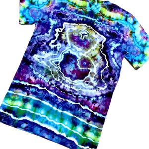 May include: A tie-dye t-shirt with a vibrant, swirling pattern in shades of blue, purple, green, and white. The design features concentric circles and wavy lines, creating a psychedelic effect. The shirt has short sleeves and a crew neck.