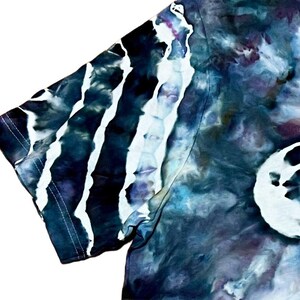 May include: A tie-dye t-shirt sleeve in shades of blue, purple, and white. The design features vertical white stripes against a dark blue background, with a marbled effect. The fabric is soft and appears to be cotton.