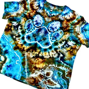 May include: A V-neck t-shirt featuring a tie-dye pattern in shades of blue, brown, and white. The center of the shirt displays a prominent white paw print graphic surrounded by swirling colors that create a marbled effect across the fabric.