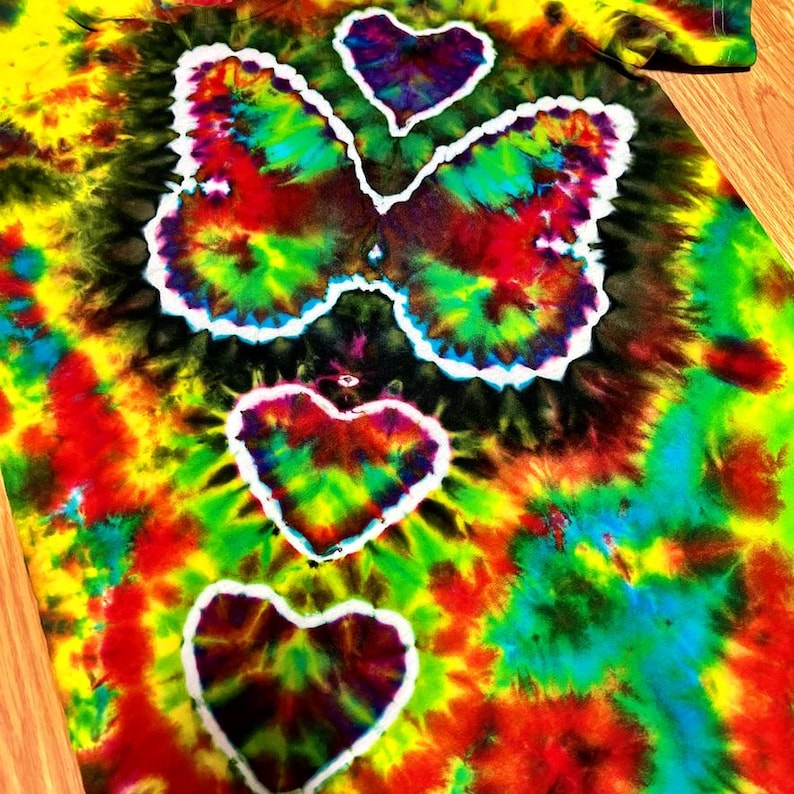 May include: A tie-dye shirt with a butterfly and heart design. The shirt features a vibrant mix of colors, including red, yellow, green, and blue. The butterfly and hearts are outlined in white, creating a striking contrast against the colorful background.