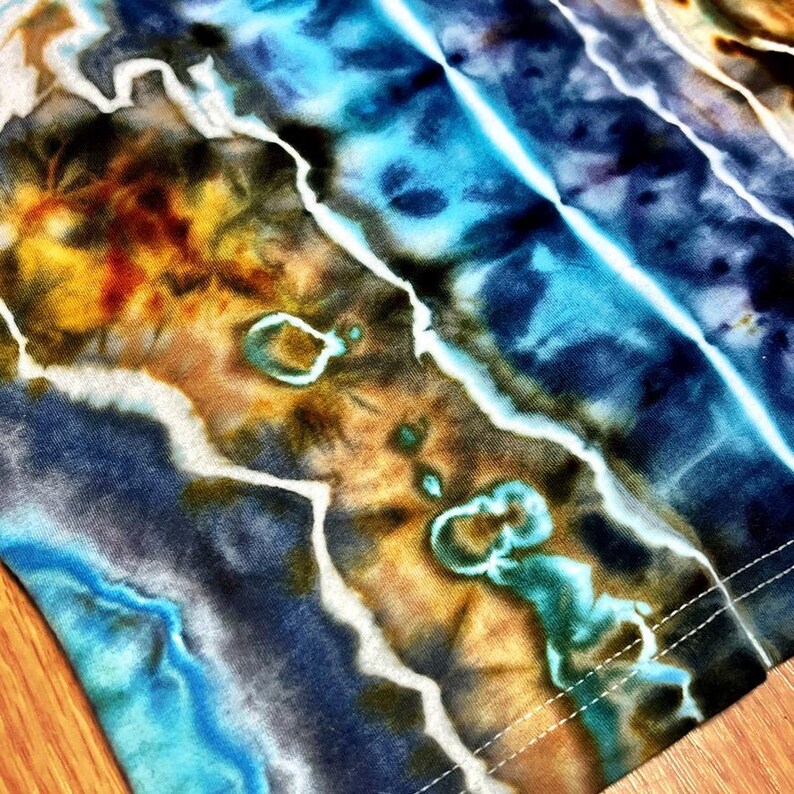 May include: Close-up of a tie-dye fabric with a blend of colors. The design features shades of blue, brown, yellow, and white, creating a marbled effect. The fabric has a soft, textured appearance with visible stitching along the edge.